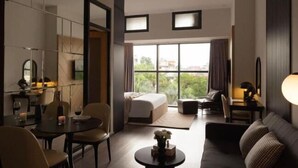 Una habitación de hotel moderna con cama, zona de estar con sofá y sillones, mesa de comedor con velas y ventanas amplias que ofrecen vistas al exterior.