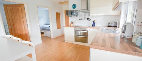 Een compacte keuken met een inbouwoven, een magnetron en een waterkoker. Er is een eethoek met een witte tafel en stoelen.