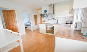 A compact kitchen with a built-in oven, a microwave, and a kettle. There is a dining area with a white table and chairs. - Swallows Croyde 3 Bedrooms, Sleeps 6, Beach Chale (Braunton)