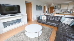 A modern living room with a flat-screen TV, a black leather sofa, a white coffee table, and a kitchen in the background. - Swallows Croyde 3 Bedrooms, Sleeps 6, Beach Chale (Braunton)
