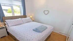 A neatly made bed with a patterned bedspread and pillows, a bedside lamp, and a heart-shaped decoration on the wall. - Swallows Croyde 3 Bedrooms, Sleeps 6, Beach Chale (Braunton)