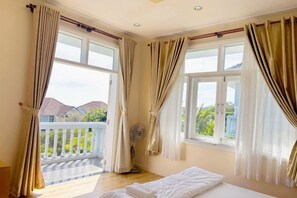 Un dormitorio con una cama, dos ventanas con cortinas, un balcón y un ventilador.