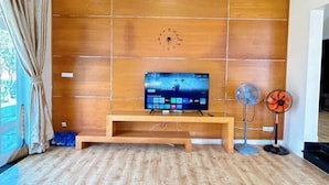 A living room with a wooden panel wall, a TV on a wooden stand, and two standing fans. - 4 bedroom Seafront Soan Villa - Sealinks Mui Ne (Phan Thiet)