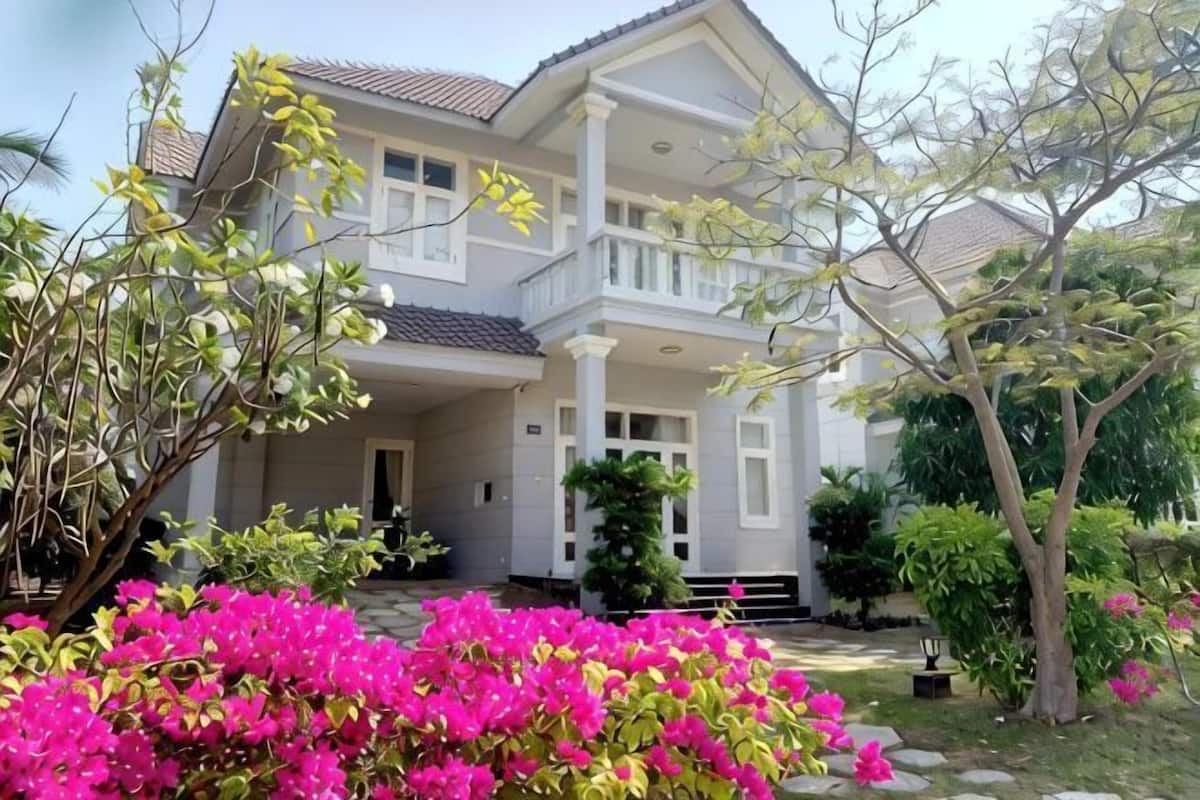 A two-story house with a balcony, surrounded by lush greenery and flowering plants.
