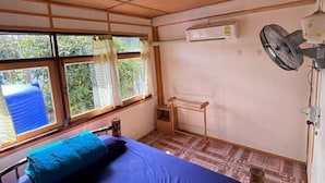 Een kamer met een bed, een airconditioning, een ventilator, een raam met gordijnen en een kleine tafel.