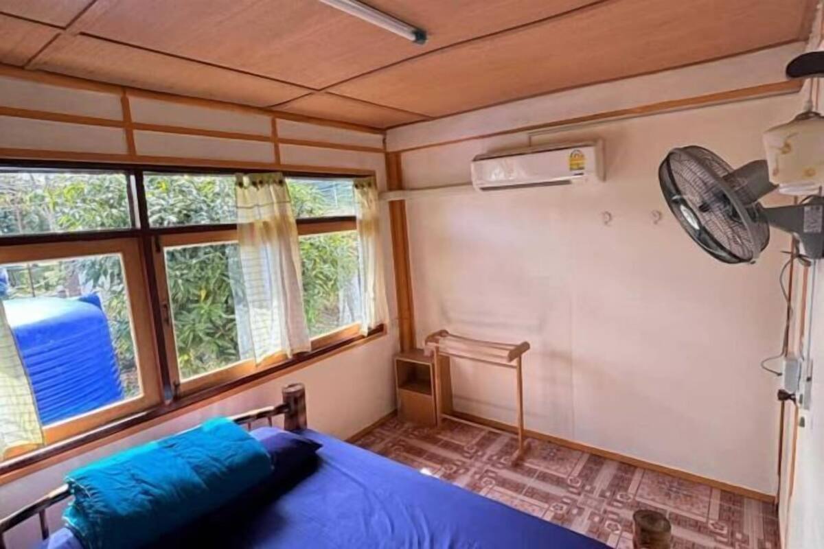 Een kamer met een bed, een airconditioning, een ventilator, een raam met gordijnen en een kleine tafel.