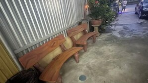 A row of wooden benches against a corrugated metal wall, a potted plant, and a concrete floor with a drain. - VietHung Hostel-Motorbikes Renta-Tuor (Ha Giang)