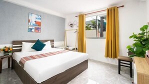 Een hotelkamer met een bed, een bureau, een stoel, een raam met gordijnen, een aan de muur hangend kunstwerk en een kleine plant.