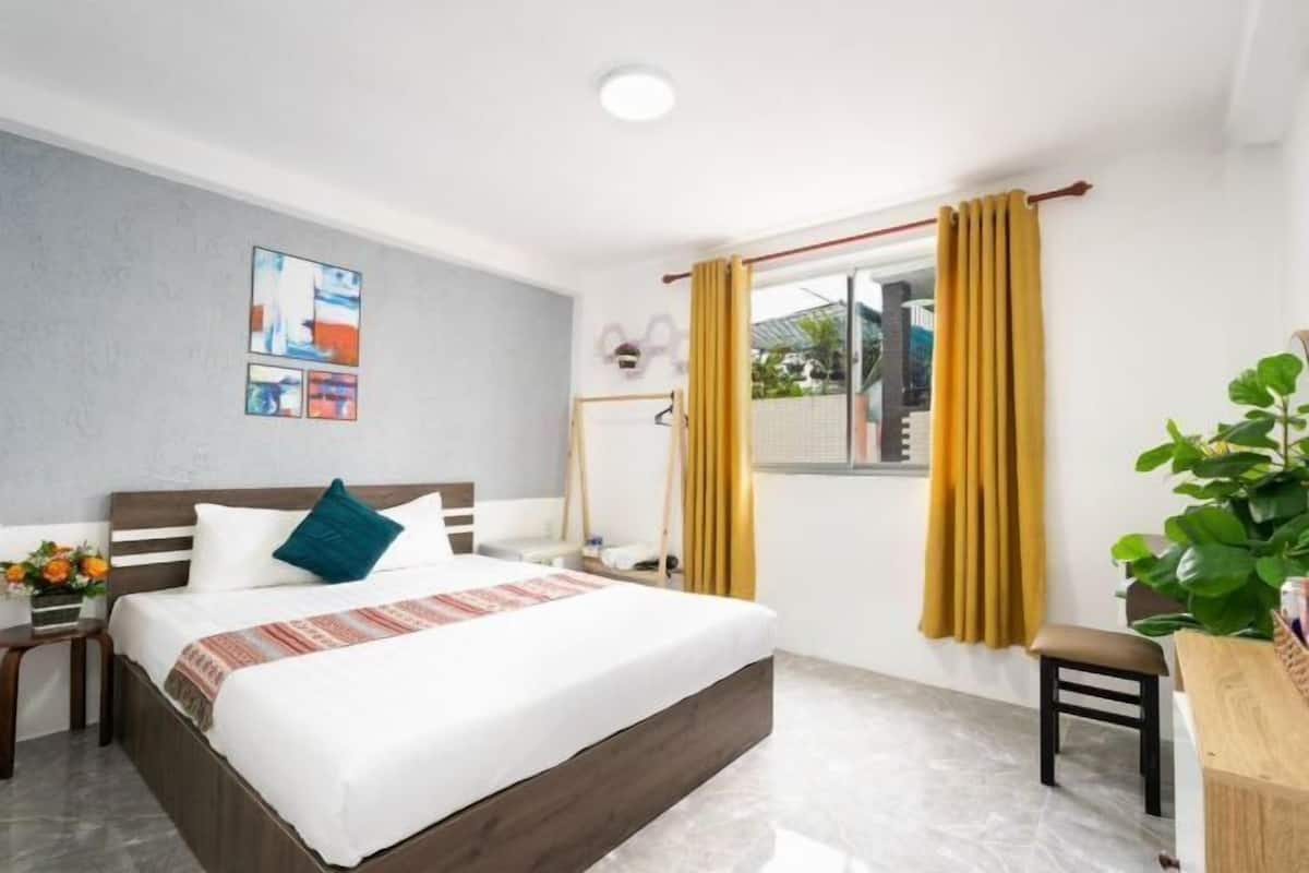Une chambre d’hôtel comprenant un lit, un bureau, une chaise, une fenêtre avec des rideaux, une œuvre d’art accrochée au mur et une petite plante.