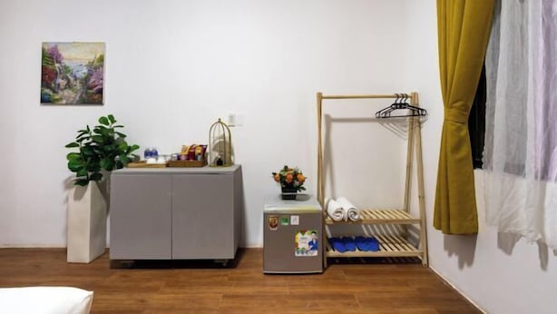 Een kamer met een bed, een klein tafeltje met een plant, een kast, een koelkast, een schoenenrek en een schilderij aan de muur.