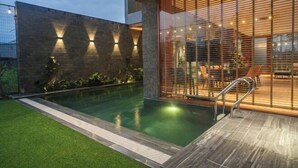 A modern outdoor pool area with a wooden deck, a stone wall with lighting, and a glass enclosure with furniture. - Villa SuSu (Da Nang)