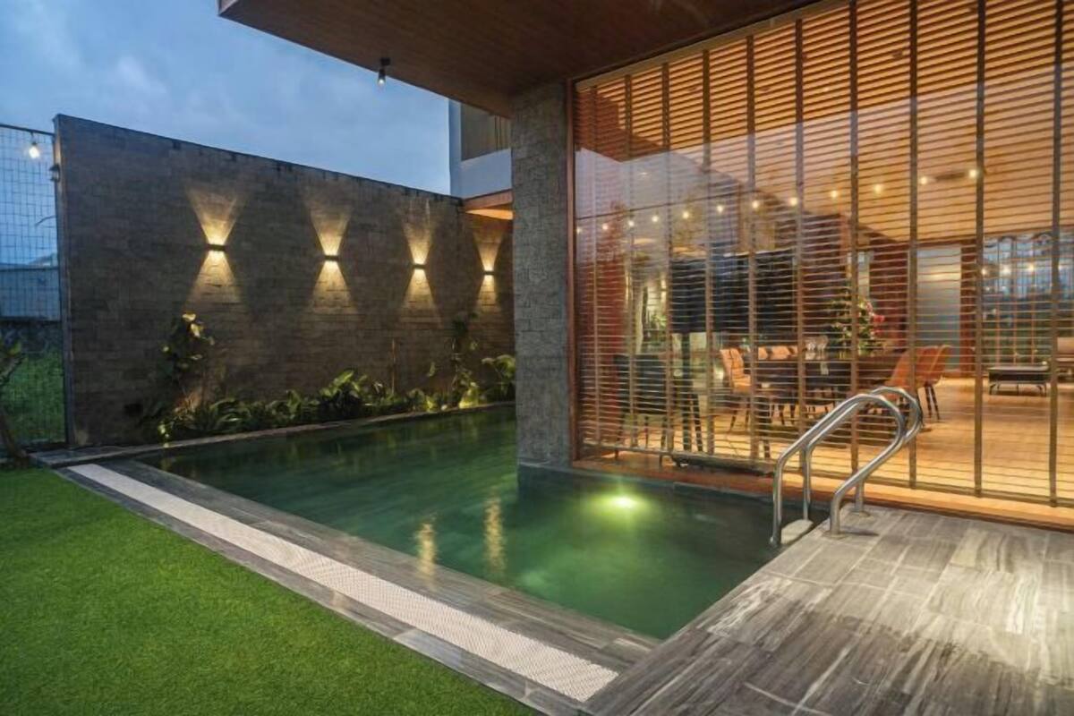 A modern outdoor pool area with a wooden deck, a stone wall with lighting, and a glass enclosure with furniture.