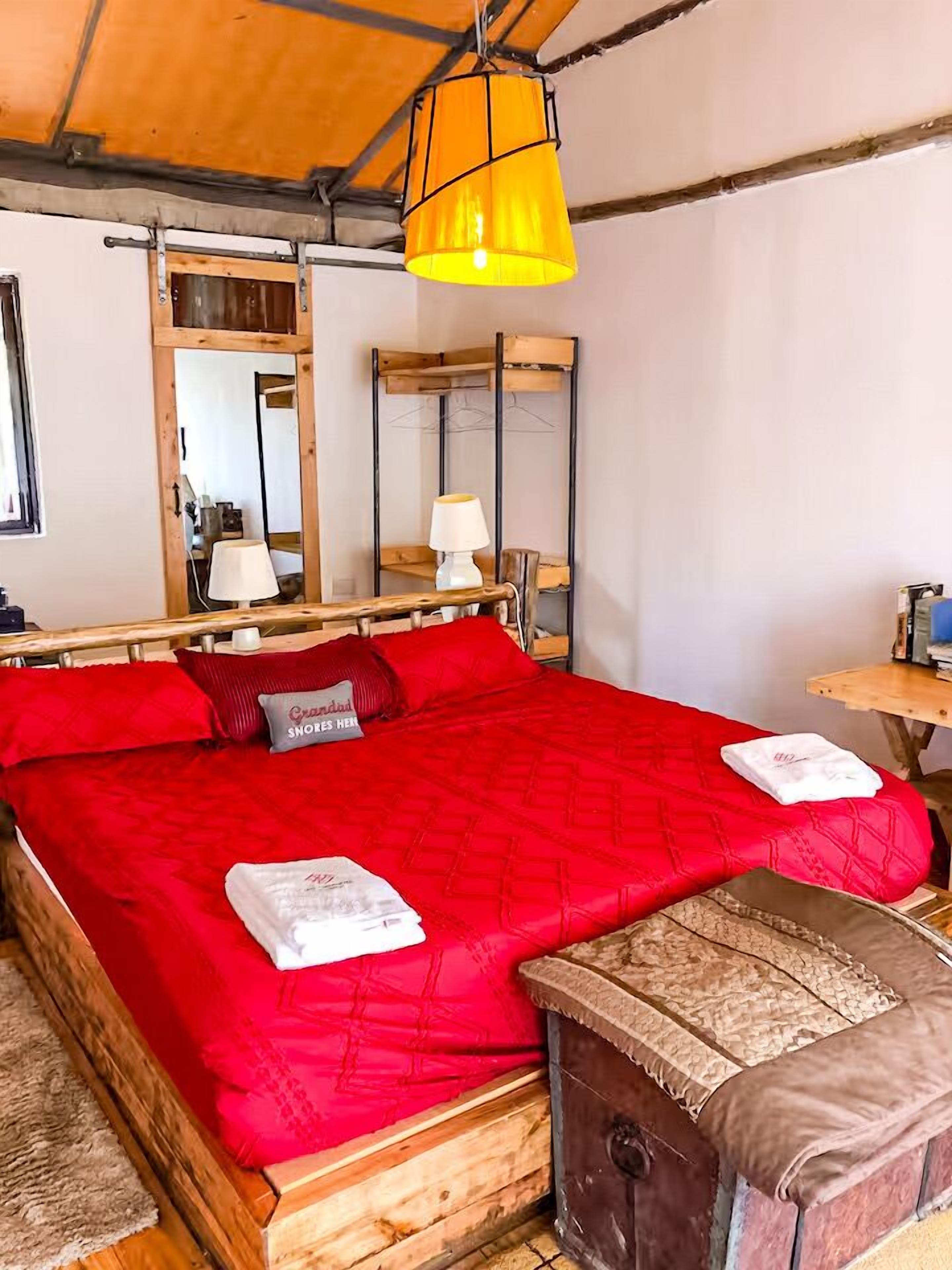 Uma cama com um edredom vermelho, um banco de madeira, uma mesinha e um abajur pendurado.