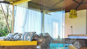 Une pièce avec un plancher en bois, un lit agrémenté d’oreillers à motifs, un carillon éolien suspendu et une grande fenêtre avec des rideaux.