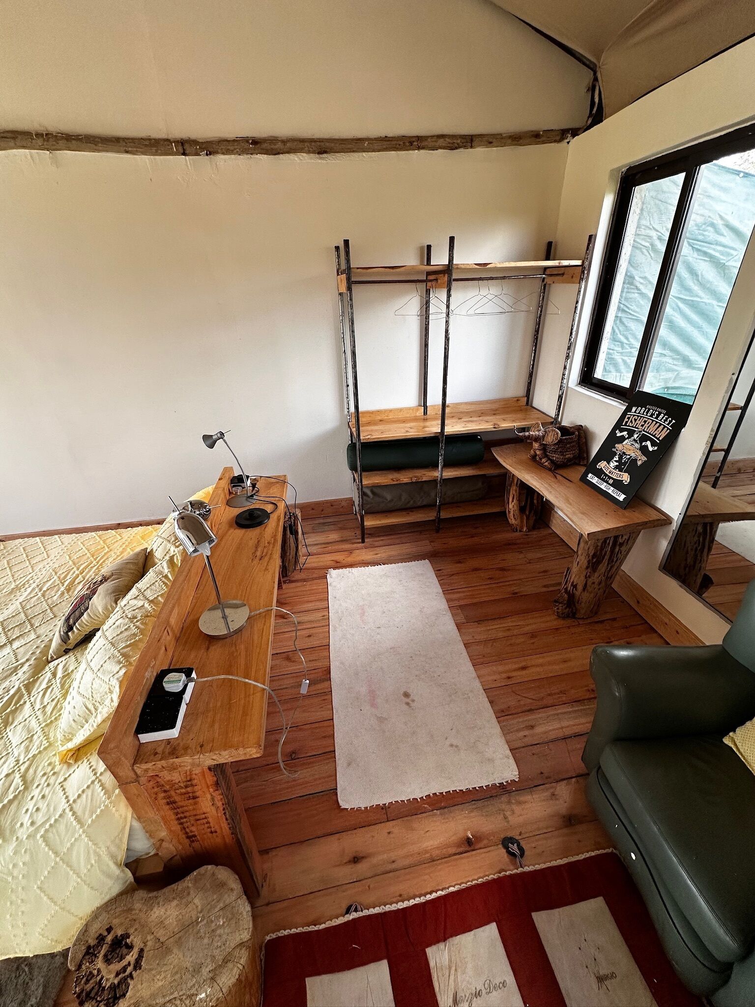 A cozy cabin interior with a wooden bench, a small table with a lamp, a hanging clothes rack, and a window with a view.