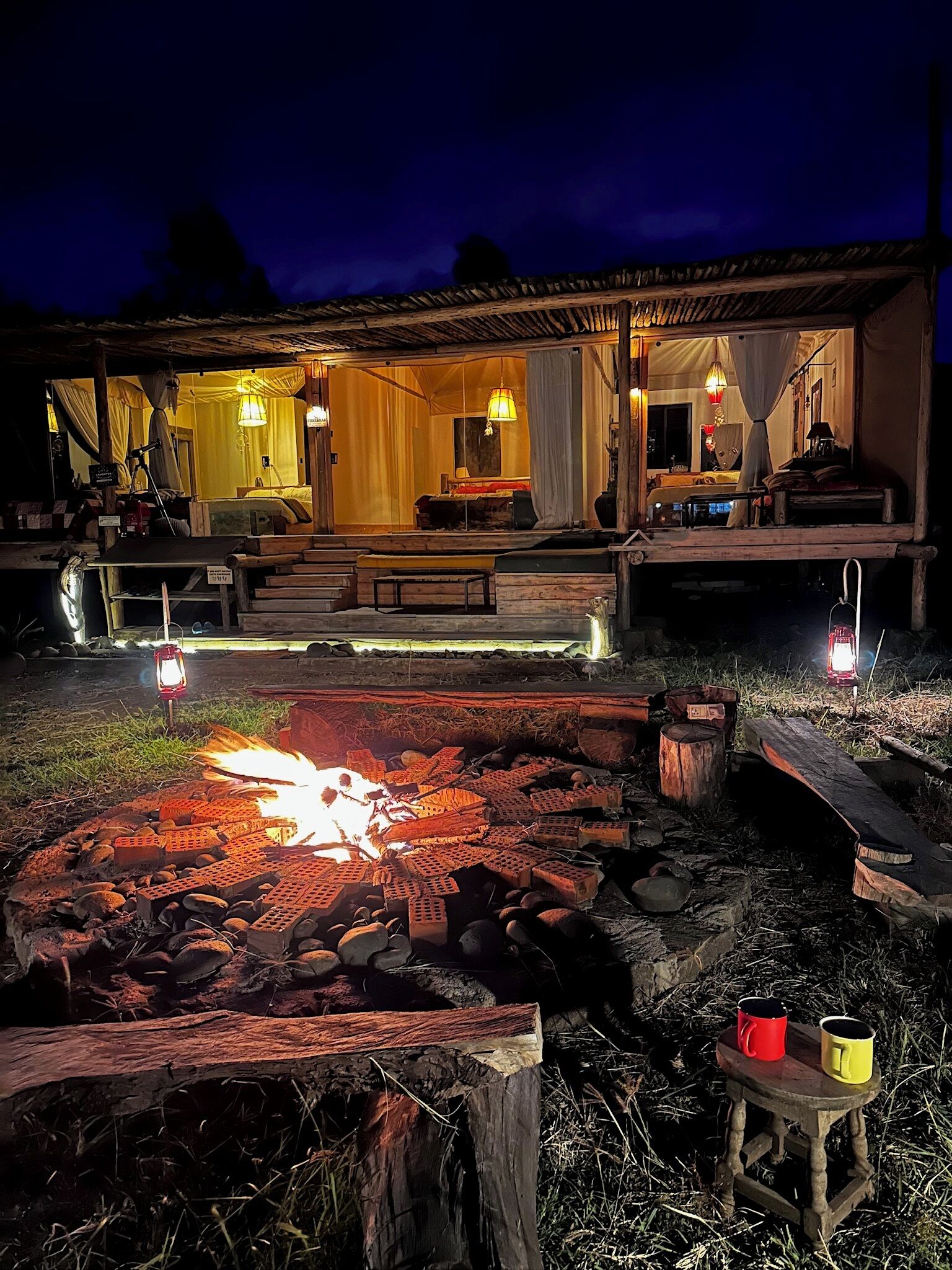 Eine rustikale Outdoor-Szenerie mit Feuerstelle, Holzbänken und einem kleinen Tisch mit Tassen.