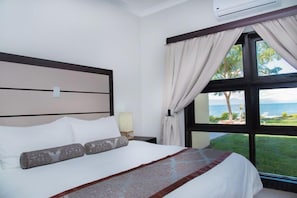 A hotel room with a large bed, a nightstand, a lamp, and a view of the ocean through large windows. - Sunbird Nkopola (Mangochi)