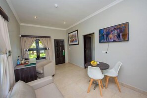 Una sala de estar con sofá, una mesa de comedor con sillas, un televisor montado en la pared y una puerta que da a otra habitación.
