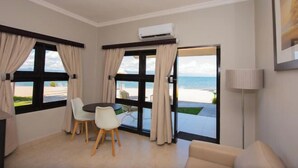 A room with a dining table and chairs, a sofa, a lamp, and large windows with a view of the beach.
