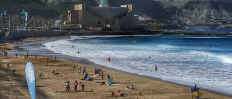 Uma praia com um prédio grande, areal arenoso e pessoas.
