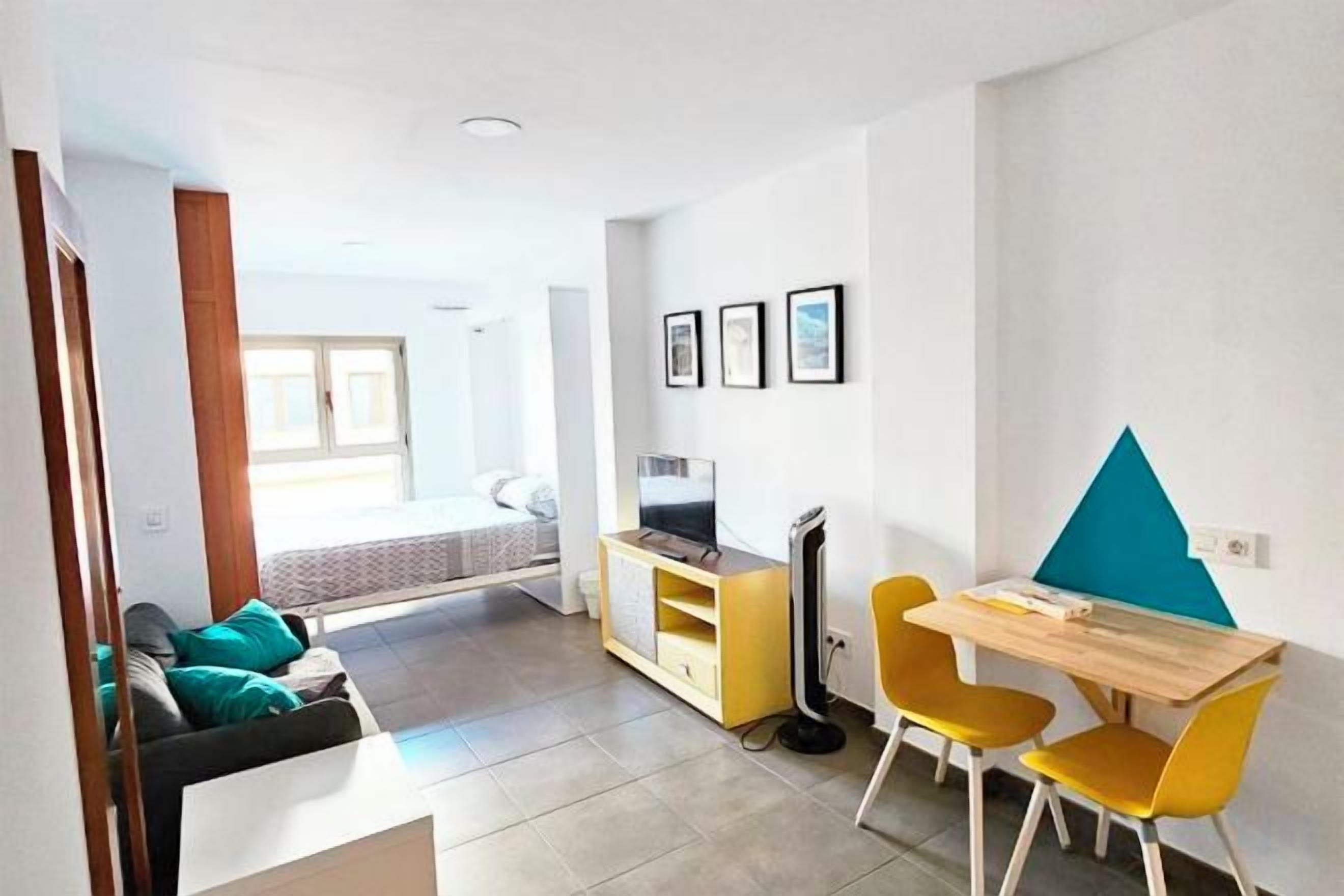 Una sala moderna con sofá, mesa de comedor con sillas, televisor y cama al fondo.