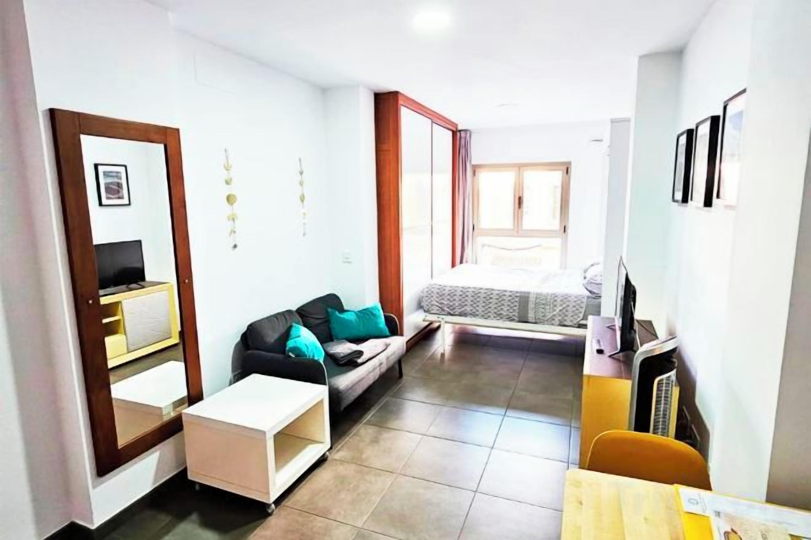 Espaço compacto para viver, com sofá, uma mesinha, TV e uma cama ao fundo.