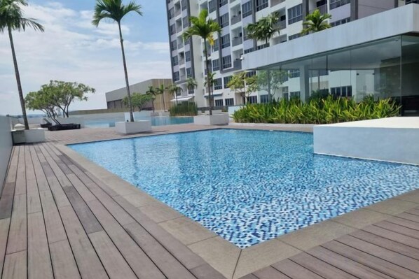 A swimming pool with a wooden deck, surrounded by modern buildings and greenery.