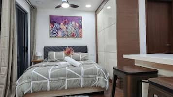 A bedroom with a bed, a desk, a chair, a ceiling fan, and a painting on the wall.