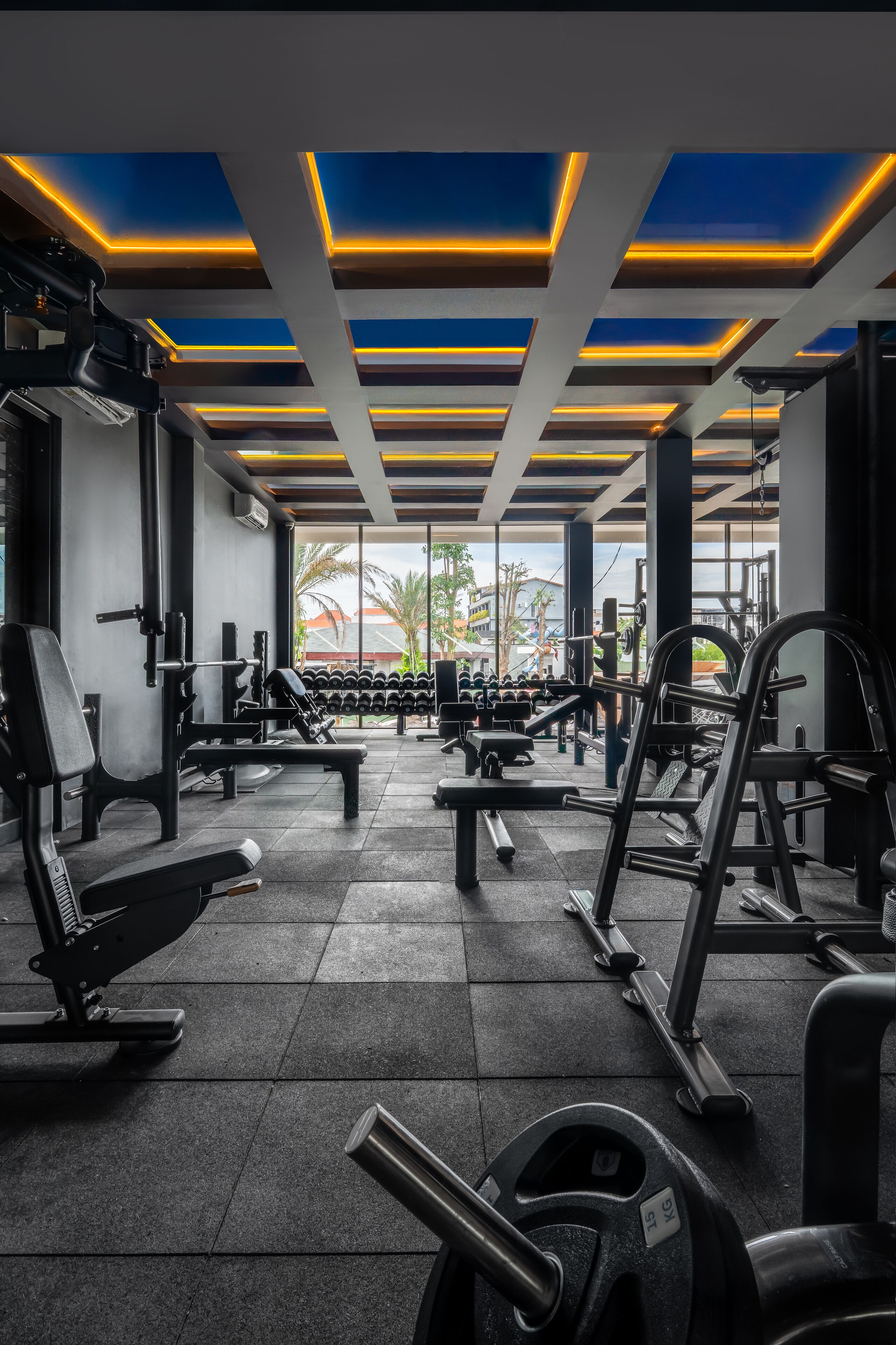 Una palestra con diverse attrezzature per esercizi, un soffitto illuminato in blu e giallo, e vista su palme all'esterno.