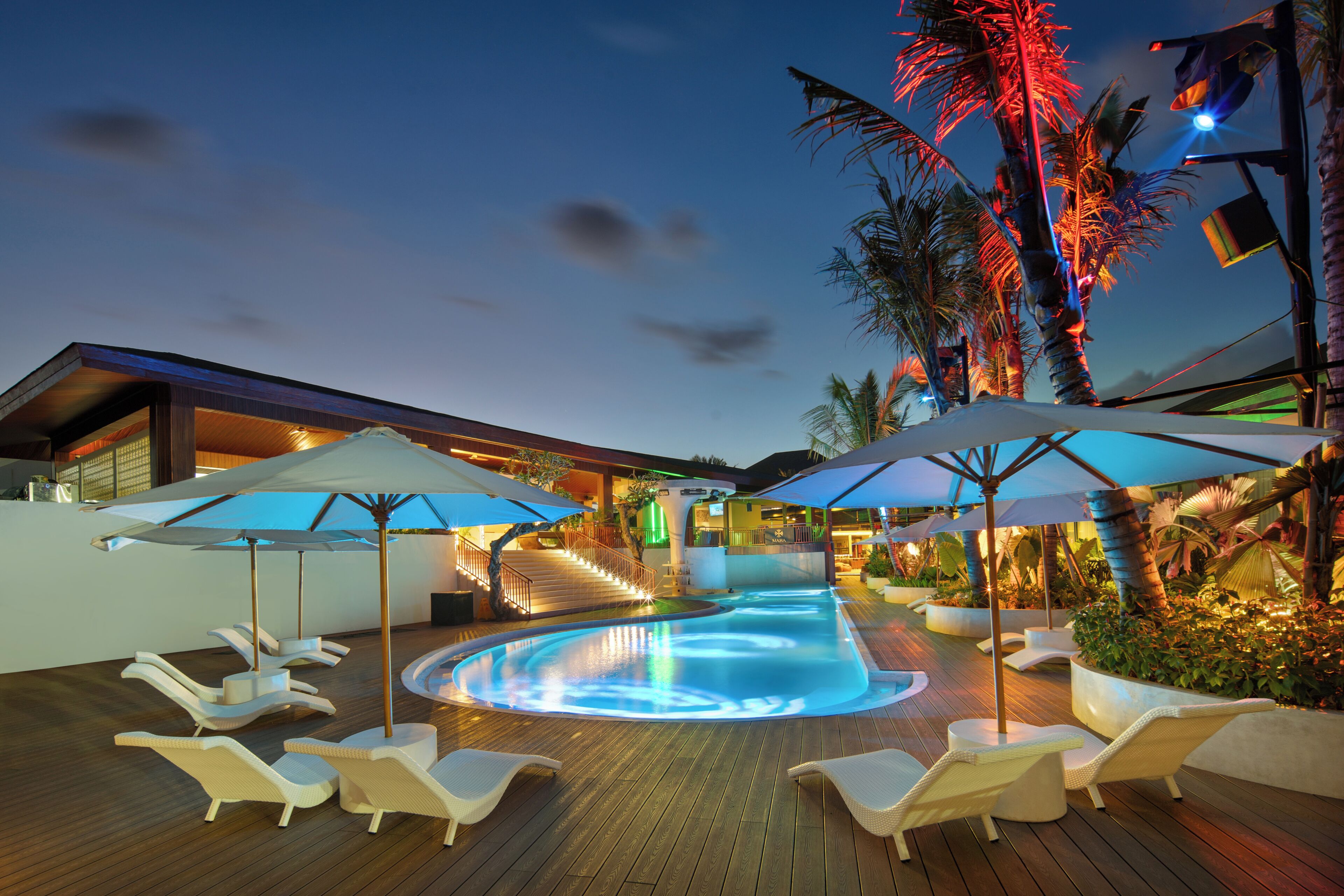 A pool area with lounge chairs and umbrellas, surrounded by palm trees and a wooden deck.
