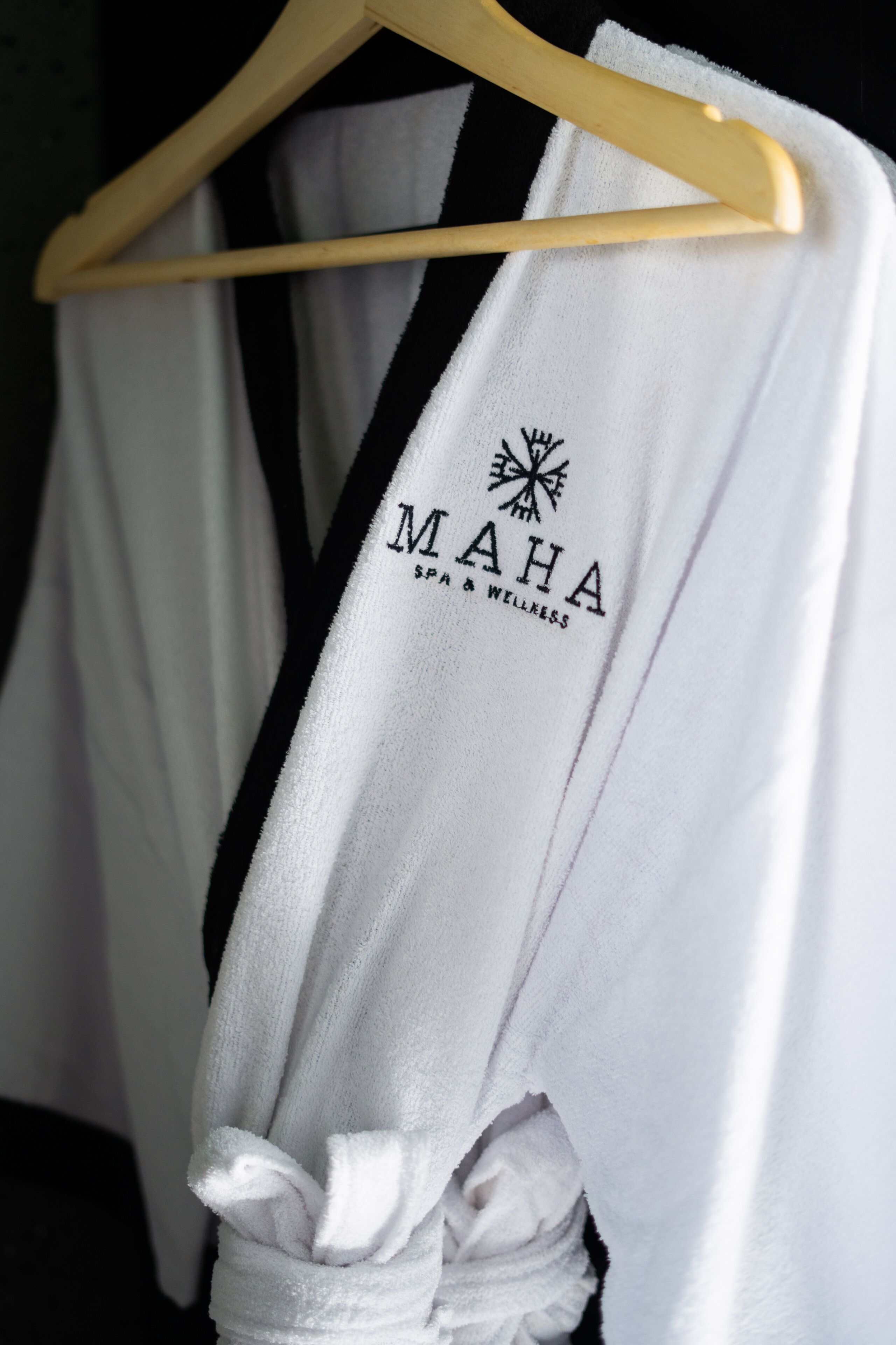 A white robe with "MAHA SPA & WELLNESS" printed on it, hanging on a wooden hanger.
