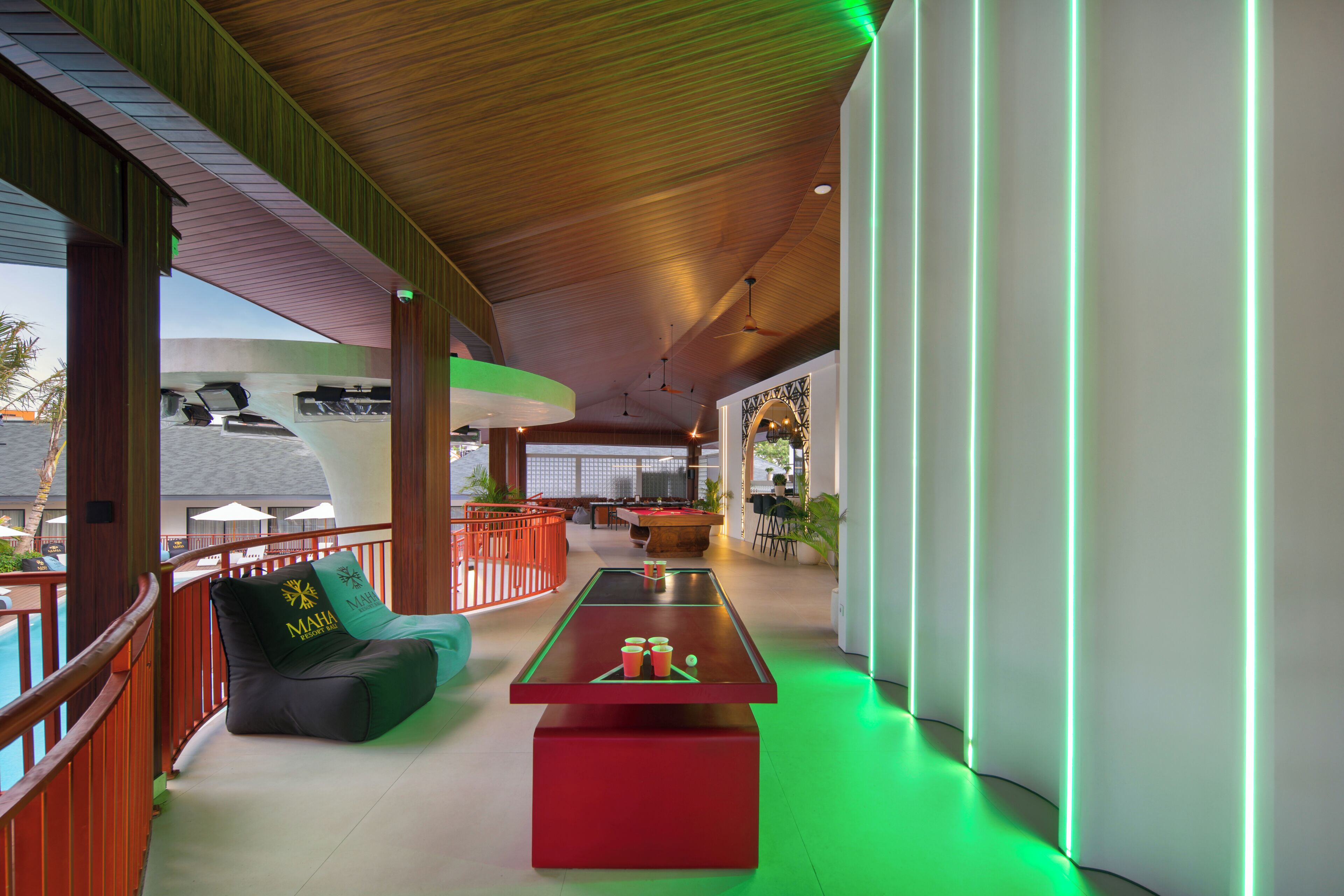 Una mesa de billar con base roja, un sillón de girable con un logotipo, y una mesa de billar en una habitación con techo de madera y vista a la piscina.