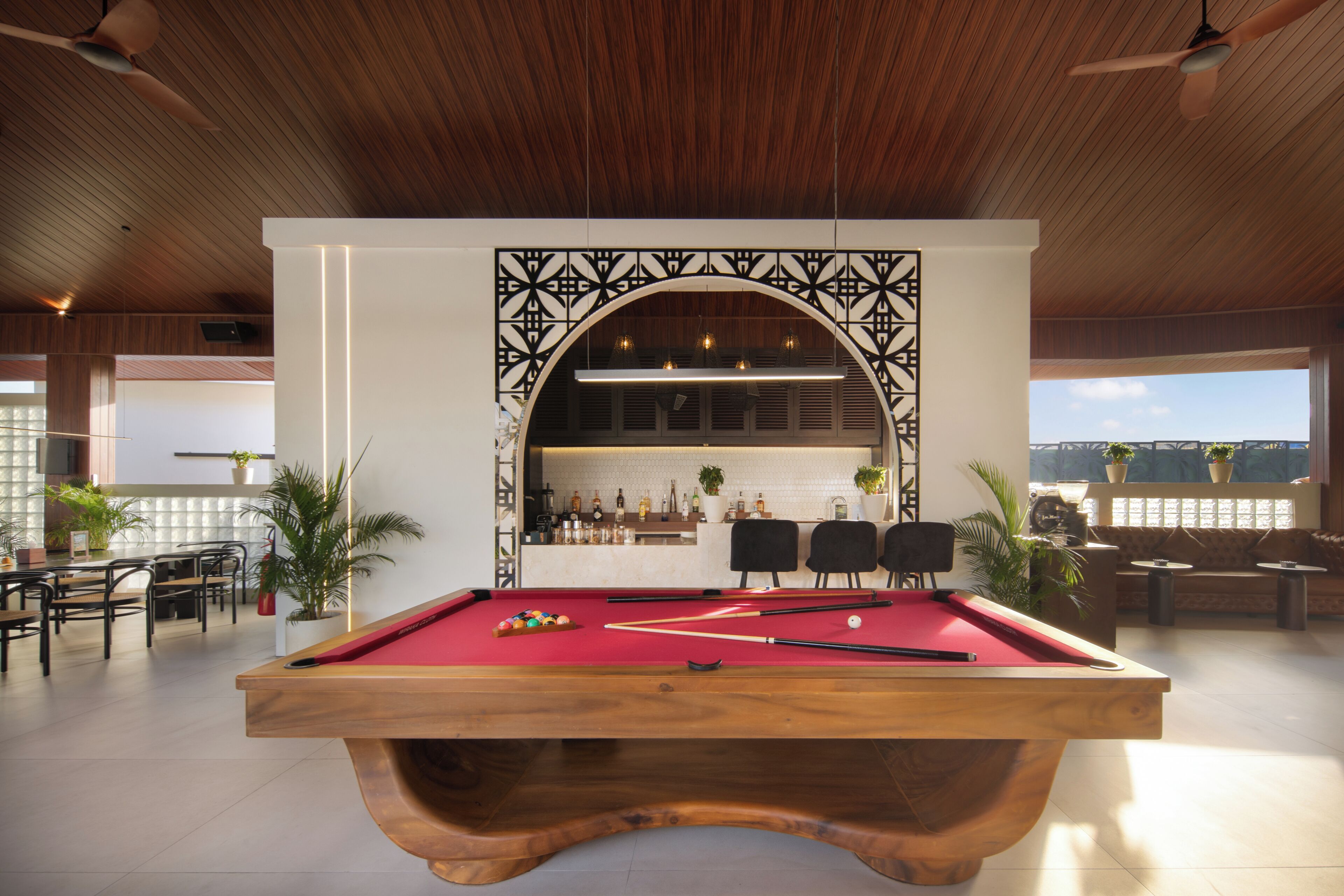 Une table de billard recouverte d’un drap rouge, entourée de chaises et d’un coin bar avec des bouteilles et des plantes.