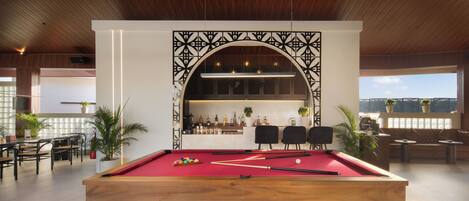 Une table de billard recouverte d’un drap rouge, entourée de chaises et d’un coin bar avec des bouteilles et des plantes.