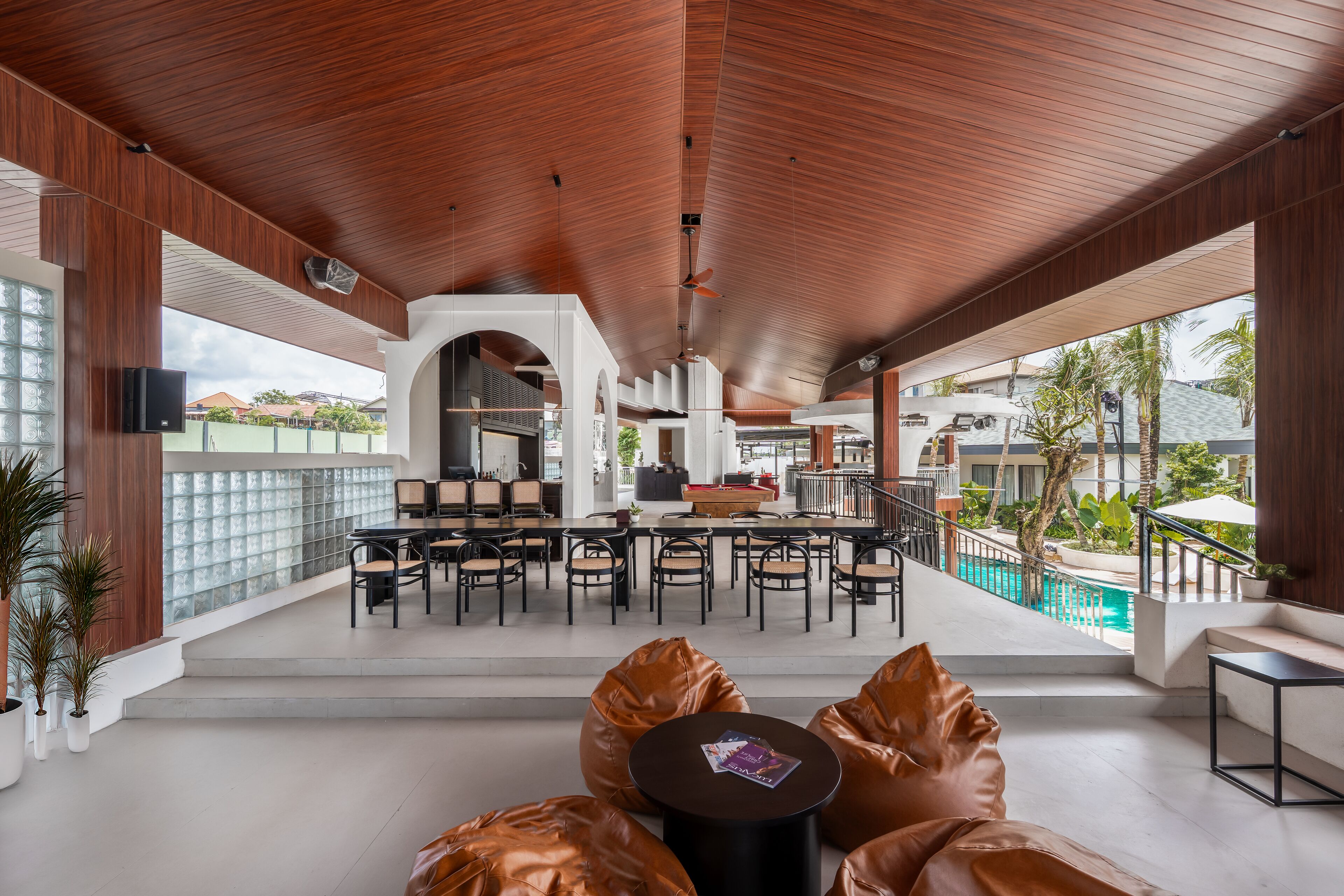 A modern outdoor lounge area with wooden ceiling, seating, and a pool view.