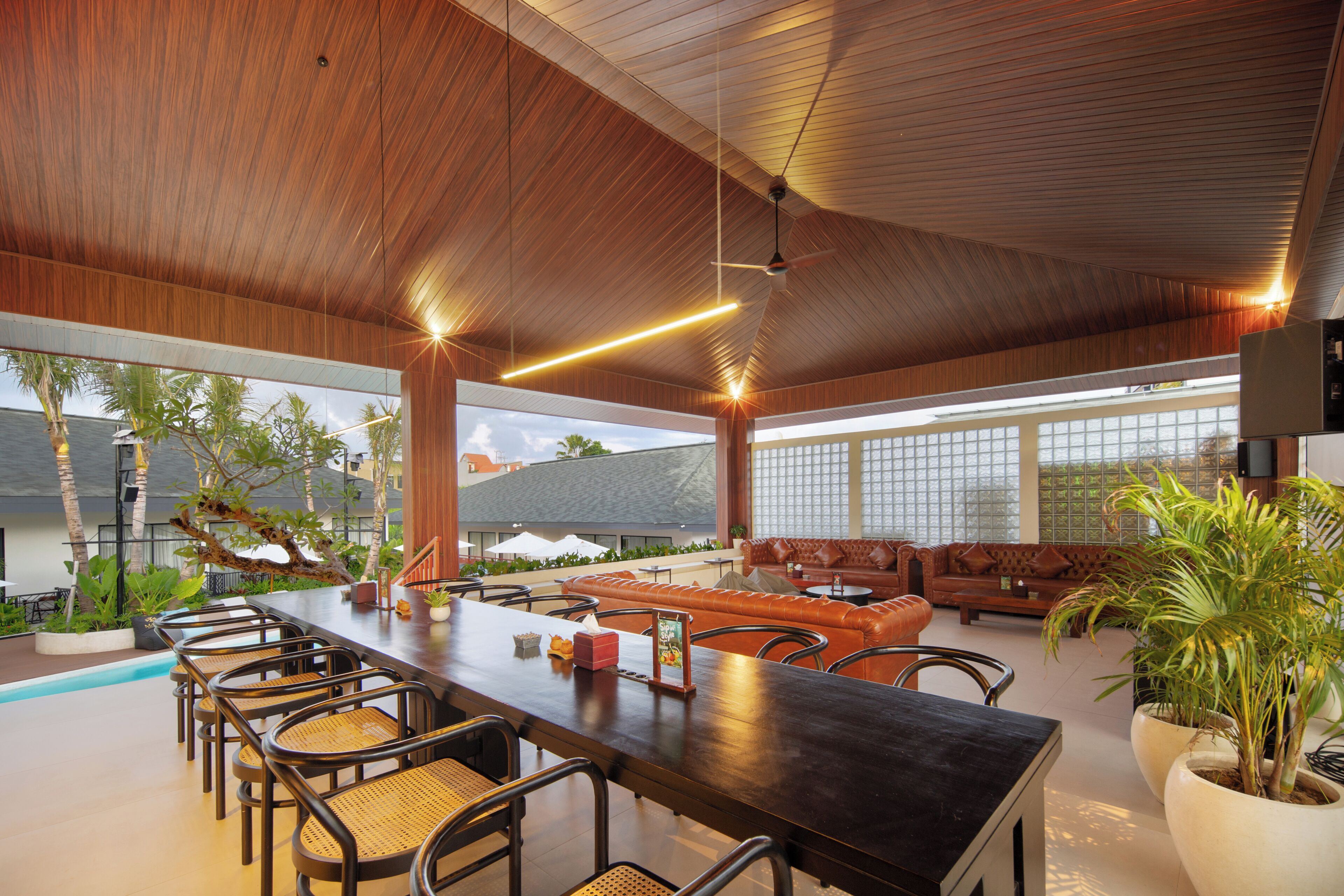 A spacious indoor area with a long wooden table, multiple chairs, and a sofa. There are large windows allowing natural light and views of the outside.