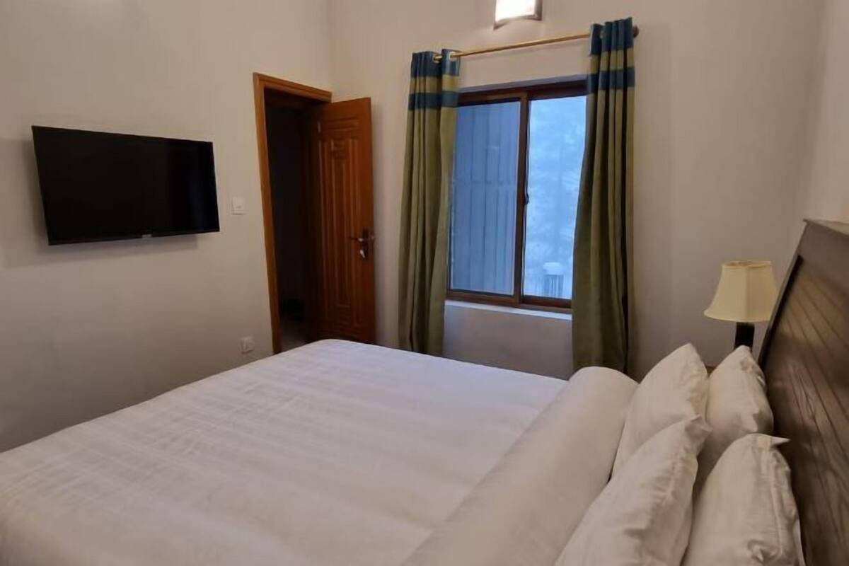 Una habitación de hotel con cama, televisión montada en la pared, ventana con cortinas y lámpara en la mesita de noche.