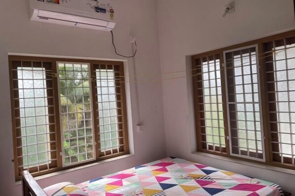 A room with a bed, two windows with metal bars, an air conditioning unit, and a view of greenery outside. - Full AC 4bhk townhouse, 10 mins from airport (Angamali)