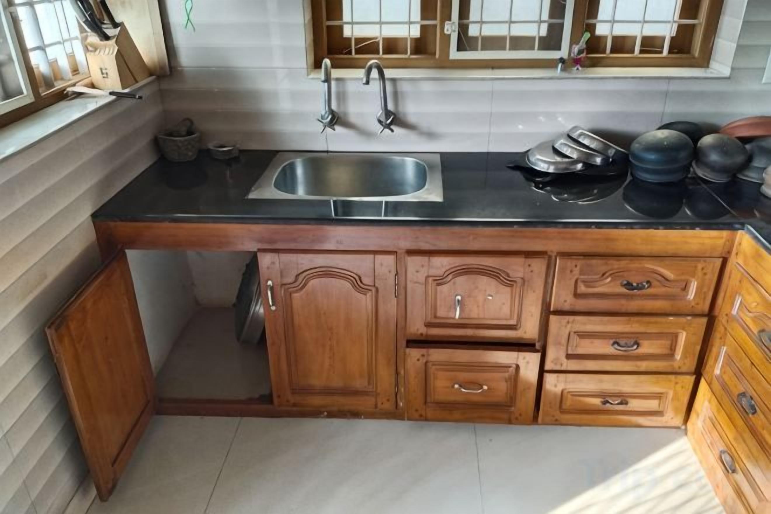 Una cocina con encimera negra, armarios de madera, un lavamanos de acero inoxidable y una ventana con diseño de cuadrícula.