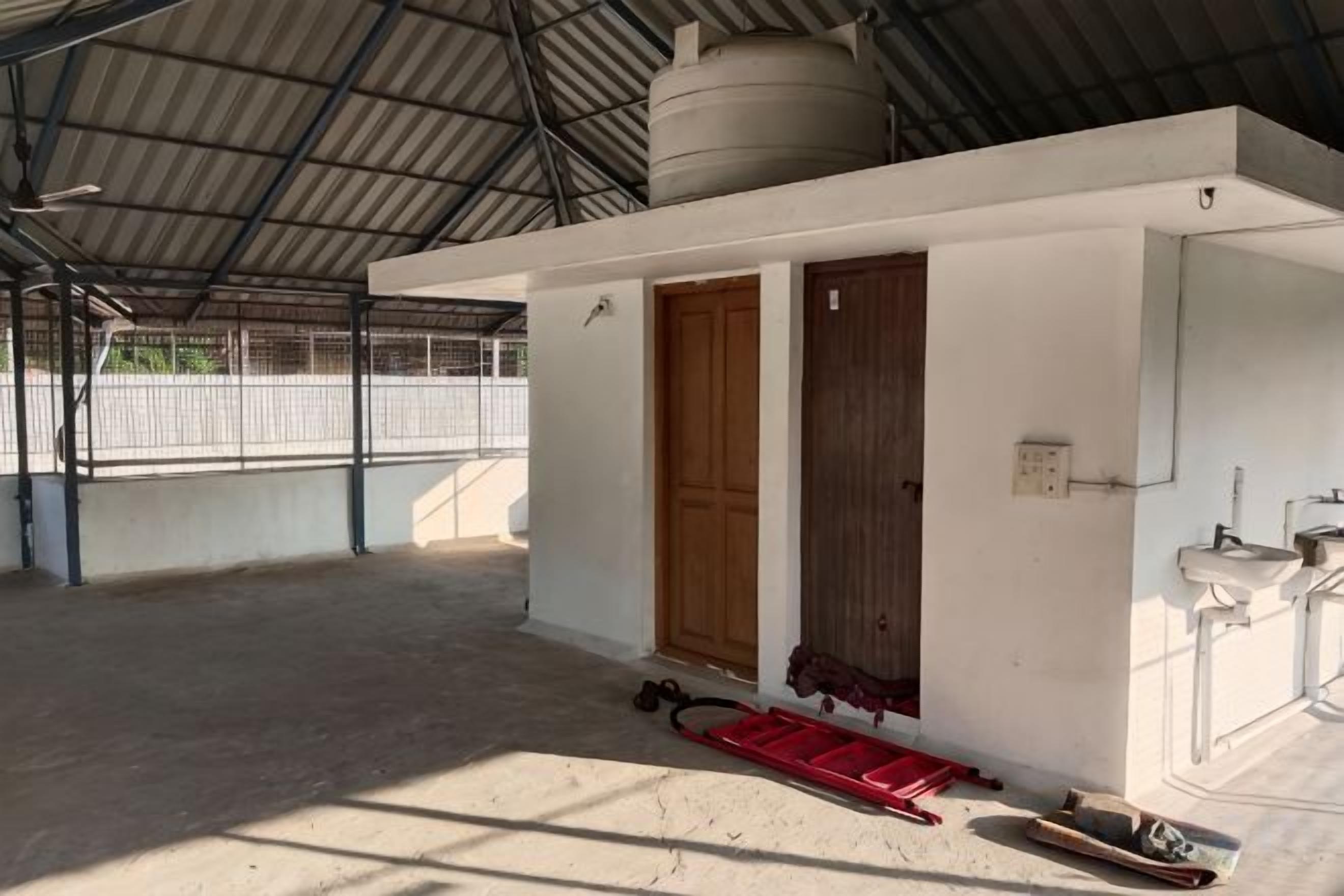 A covered outdoor area with a white building, two wooden doors, a red object on the floor, and a large water tank on the roof.