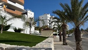 A modern building with a curved facade, a stone retaining wall, and palm trees lining the sidewalk. - Luxurious 2-Bedroom Apartment in Arenals del Sol with Pool and Sea Views (Arenals del Sol)