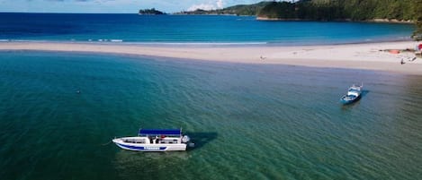 A boat on clear turquoise water near a sandy beach with a forested island in the background.