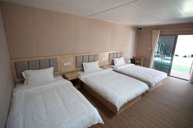A hotel room with three beds, wooden flooring, and a view of the outdoors.