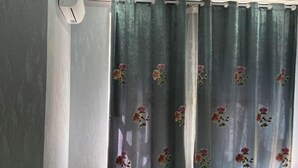 A room with a grey wall, a window with a green curtain featuring floral patterns, and a white air conditioning unit mounted on the wall. - Ali Boutique Guesthouse (Almaty)