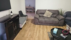 A living room with a sofa, a TV, a small table with a decorative item, and a wall clock. - Charming 3-Bedroom House in Meadow Heights - Perfect Family Retreat (Meadow Heights)