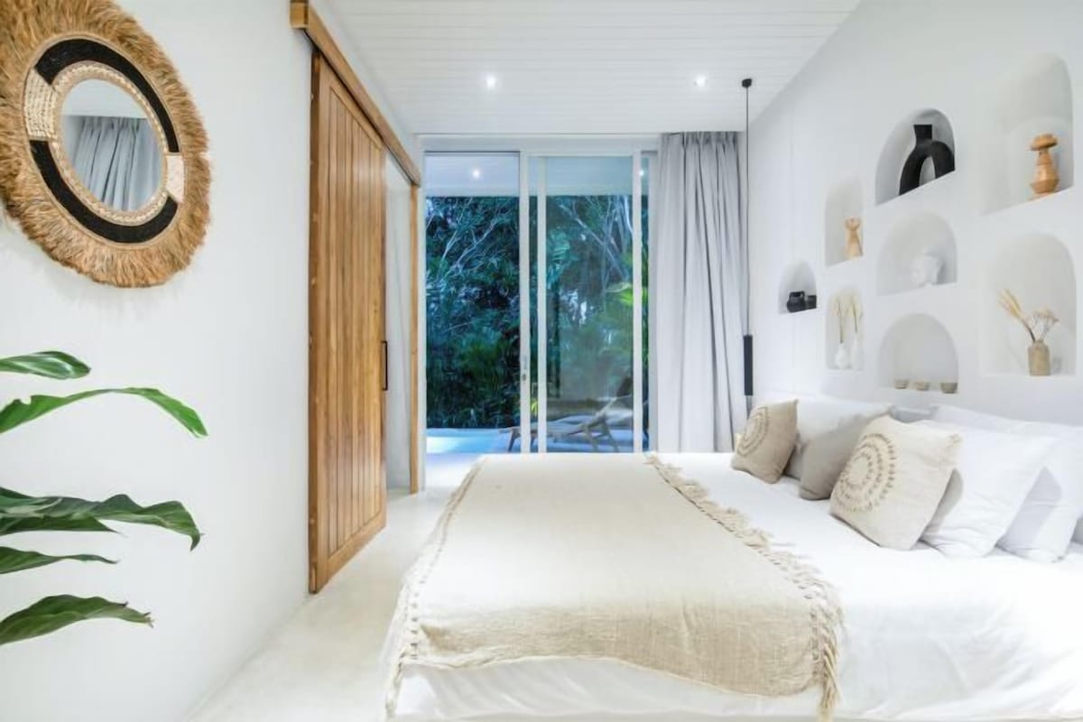 A bedroom with a bed, a large mirror, a plant, and a view of the outdoors.