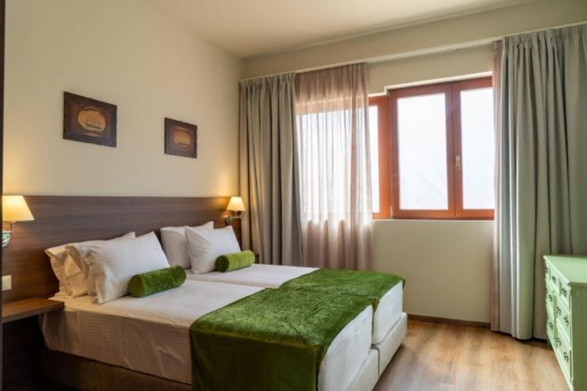 Une chambre d’hôtel comprenant un lit, deux tables de chevet, une commode et une fenêtre avec des rideaux.