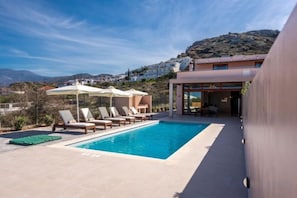 Une piscine avec des chaises longues et des parasols, un bâtiment doté d’une porte en verre, et une colline en arrière-plan.
