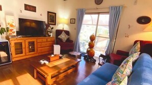 Uma sala de estar com televisão, móveis de madeira, sofá e janela com cortinas.