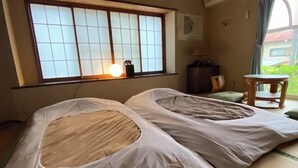 Quarto com esteiras de tatame, cama futon, janela com portas shoji, uma pequena mesa e uma cadeira.